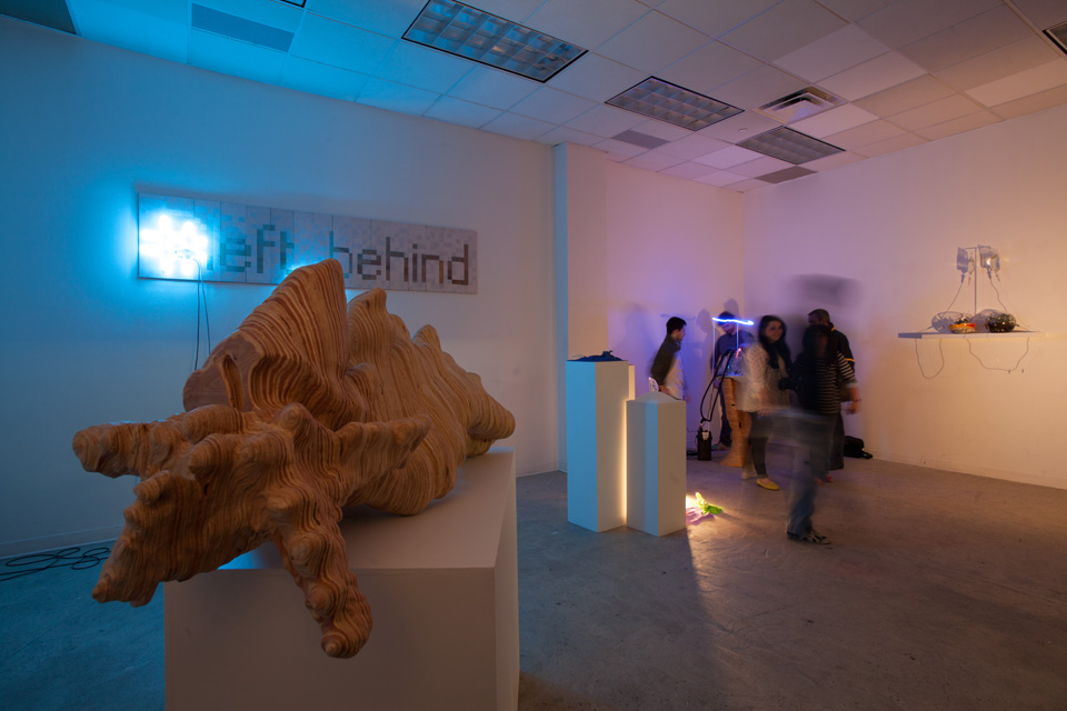 Exhibition view of bio art with organic-shaped wood sculpture, blue neon and several glass objects